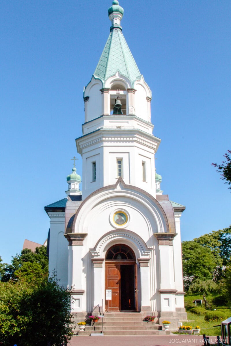 A white Orthodox church with a green, pointed dome and arched doorway stands under a clear blue sky in Hakodate Motomachi District, surrounded by greenery and small bushes.