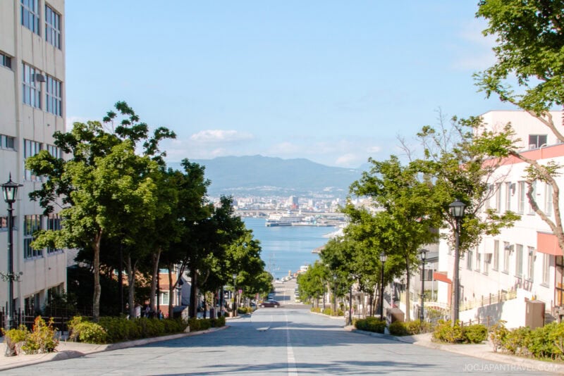 A wide, sloping street lined with trees and buildings in the Hakodate Motomachi District leads down to a distant harbor, framed by mountains and a clear blue sky.