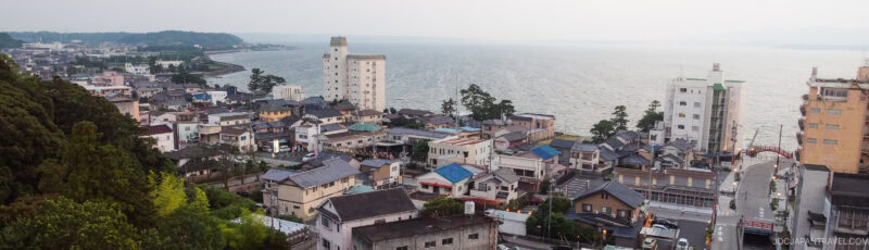 A coastal town with houses and low-rise buildings sits beside a large body of water, with a few taller buildings and a tree-covered hill visible on the left. The scene appears calm and slightly overcast.
