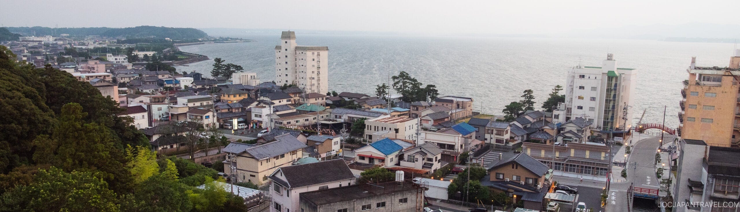 A coastal town with houses and low-rise buildings sits beside a large body of water, with a few taller buildings and a tree-covered hill visible on the left. The scene appears calm and slightly overcast.