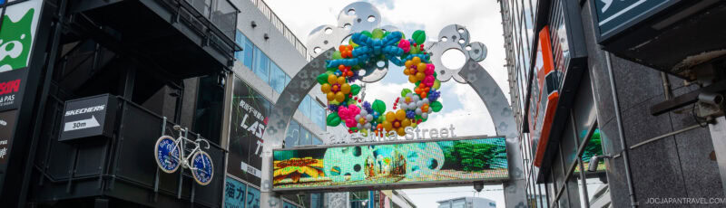 A colorful balloon wreath decorates the entrance arch of Takeshita Street in Tokyo, with a digital screen below displaying ocean-themed images and buildings on either side.