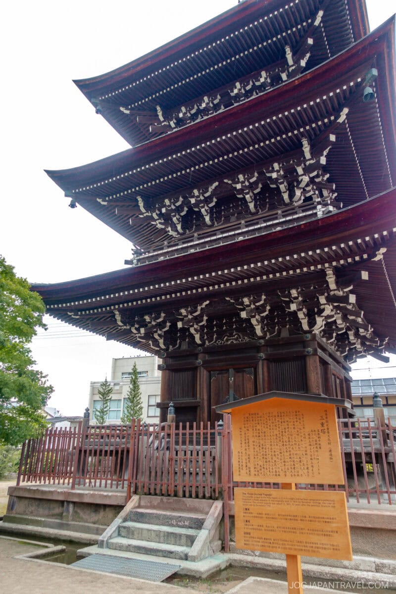 A three-story wooden pagoda with intricate eaves stands at Hida Kokubun-ji Temple, surrounded by a fence and a large orange information sign in front. Trees and buildings can be seen in the background.
