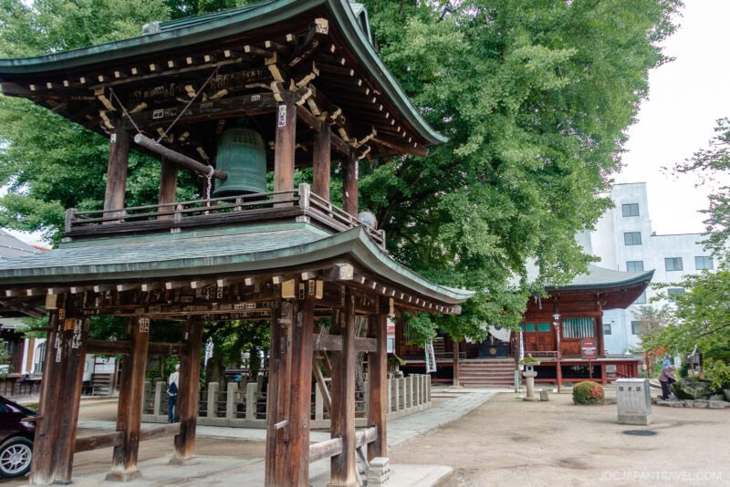 A traditional Japanese temple courtyard at Hida Kokubun-ji Temple with a large wooden bell tower in the foreground, trees, stone lanterns, and a temple building in the background. Few people are seen walking and sitting around the quiet area.