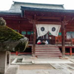 Hida Kokubun-ji Temple (飛騨国分寺) – Ancient Temple and Ginkgo Tree in Takayama A traditional wooden Hida Kokubun-ji Temple features a white cloth with black emblems at the entrance. Two people sit on the steps, while a moss-covered stone lantern stands in the foreground.