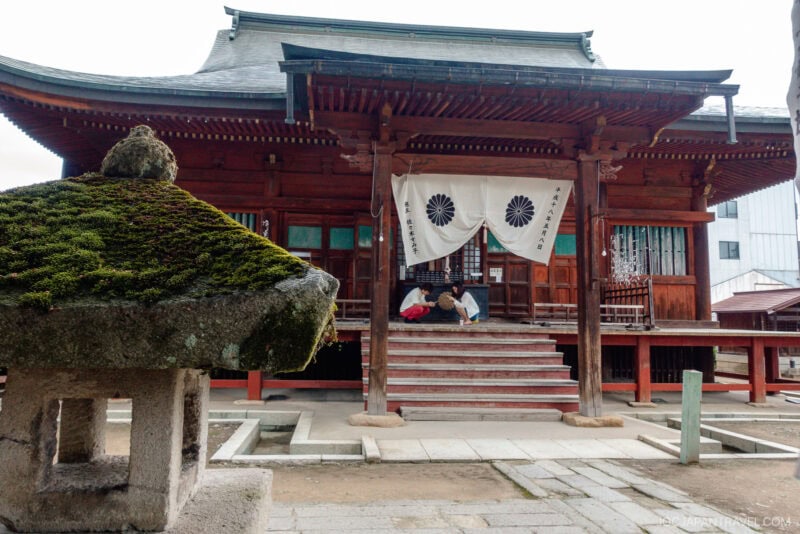 A traditional wooden Hida Kokubun-ji Temple features a white cloth with black emblems at the entrance. Two people sit on the steps, while a moss-covered stone lantern stands in the foreground.