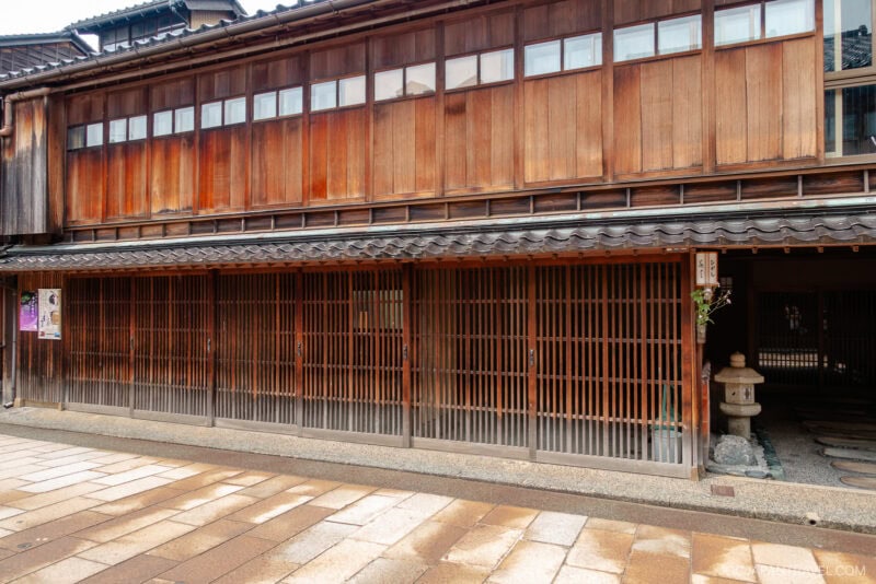 A traditional Japanese wooden building in Higashi Chaya with sliding lattice doors, tiled roof, and upper windows, located on a paved street with a stone lantern near the entrance.