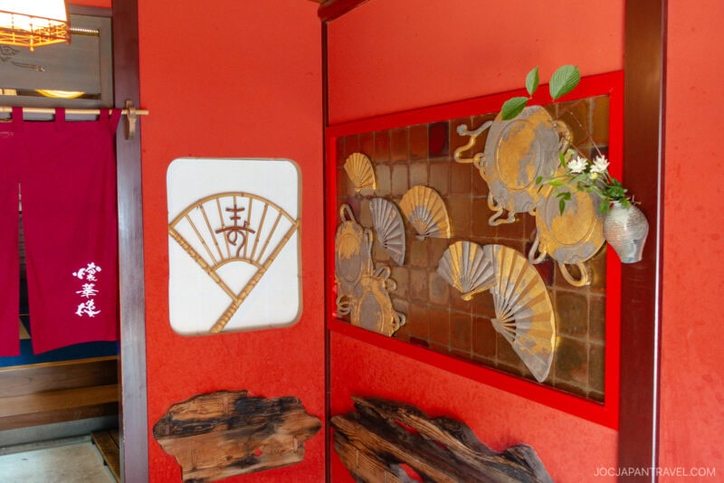A vibrant red entryway at Higashi Chaya features decorative fans and crabs on the wall, a wooden accent piece, a small vase with flowers, and red noren curtains adorned with white Japanese characters.