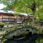 A traditional Japanese temple with wooden architecture stands behind a small stone bridge over a pond, surrounded by lush green trees and moss-covered rocks along the scenic Higashiyama Walking Course in a peaceful garden setting.