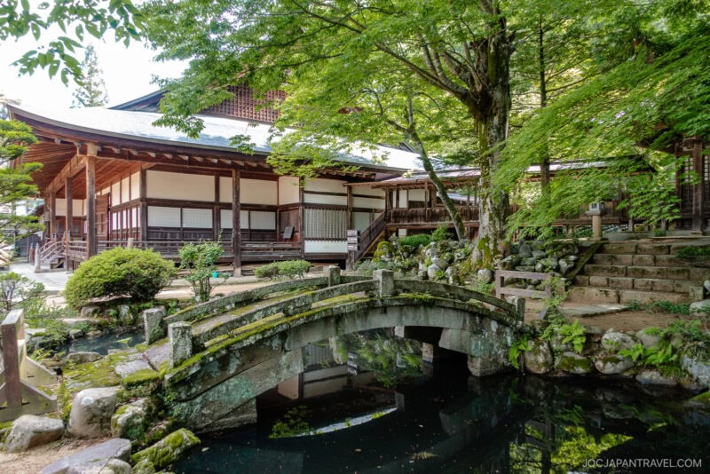 A traditional Japanese temple with wooden architecture stands behind a small stone bridge over a pond, surrounded by lush green trees and moss-covered rocks along the scenic Higashiyama Walking Course in a peaceful garden setting.