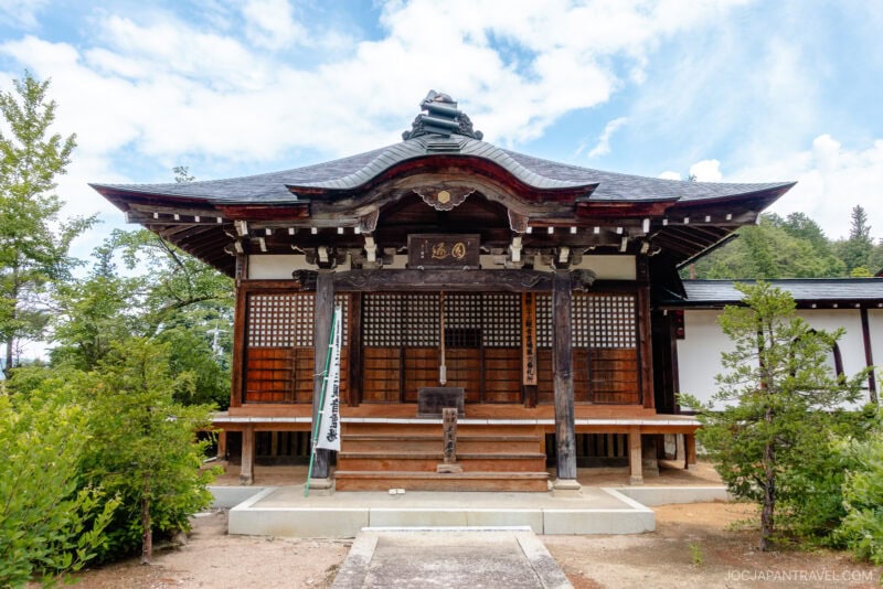 A traditional Japanese wooden temple with ornate roof details, set amidst greenery under a blue sky with scattered clouds. A small path, part of the Higashiyama Walking Course, leads to the entrance steps.