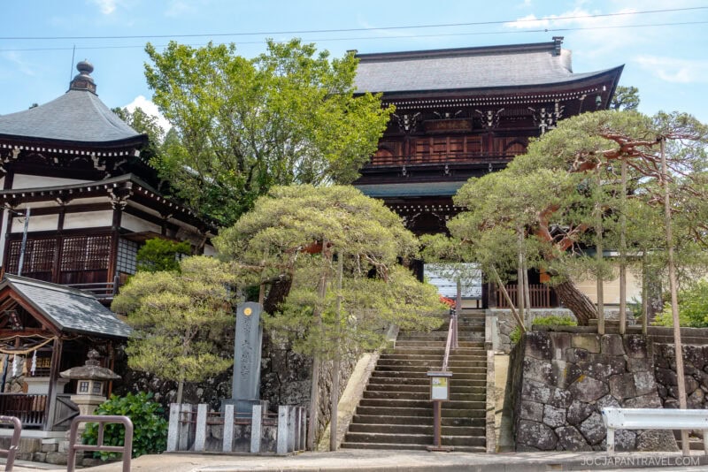 A traditional Japanese temple with wooden structures, stone steps leading up, and manicured trees and shrubs on either side, set along the scenic Higashiyama Walking Course under a bright blue sky.