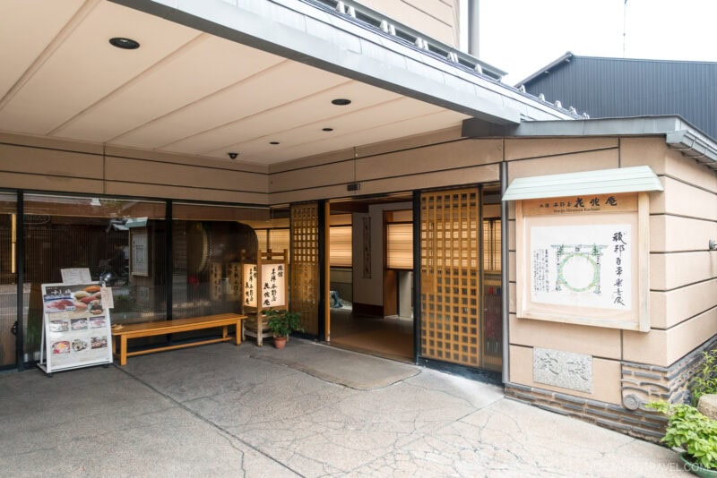 The entrance of Honjin Hiranoya Kachoan Takayama features beige walls, sliding wooden doors, a signboard, a bench, and a potted plant near the doorway.