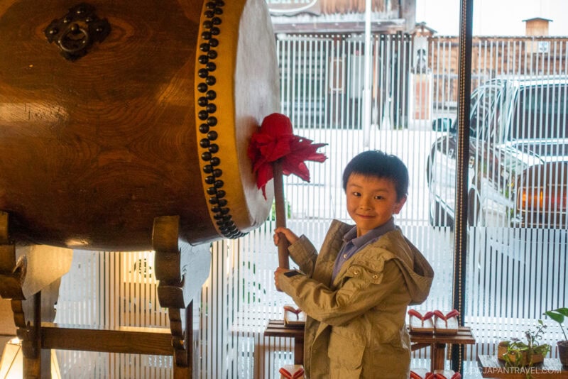 A young boy smiles while holding a red drumstick, about to hit a large traditional Japanese drum inside the Honjin Hiranoya Kachoan Takayama, with glass windows and light streaming in.