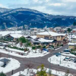 Soak in Serenity at Hyakurakusou – Yuwaku Onsen’s Hidden Gem A snowy Japanese village, featuring houses, parked cars, and trees, nestled among snow-covered mountains beneath a cloudy sky.