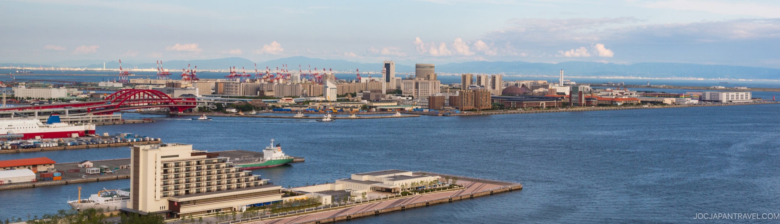 A panoramic view of a harbor cityscape with modern buildings, bridges, and cranes along the waterfront under a partly cloudy sky, with calm blue water in the foreground.