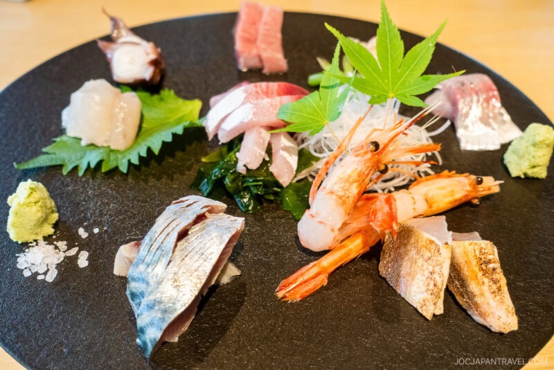At Izakaya Dai Toyama, a black plate with assorted sashimi&mdash;shrimp, fish, and octopus&mdash;is garnished with a green maple leaf, shiso leaf, wasabi, and salt, all artfully arranged on a wooden table.