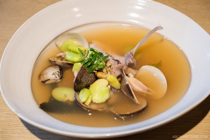A white bowl filled with clear broth, clams, broad beans, and a fish piece with bone, garnished with fresh herbs—an elegant dish at Izakaya Dai Toyama, placed on a light wooden table.