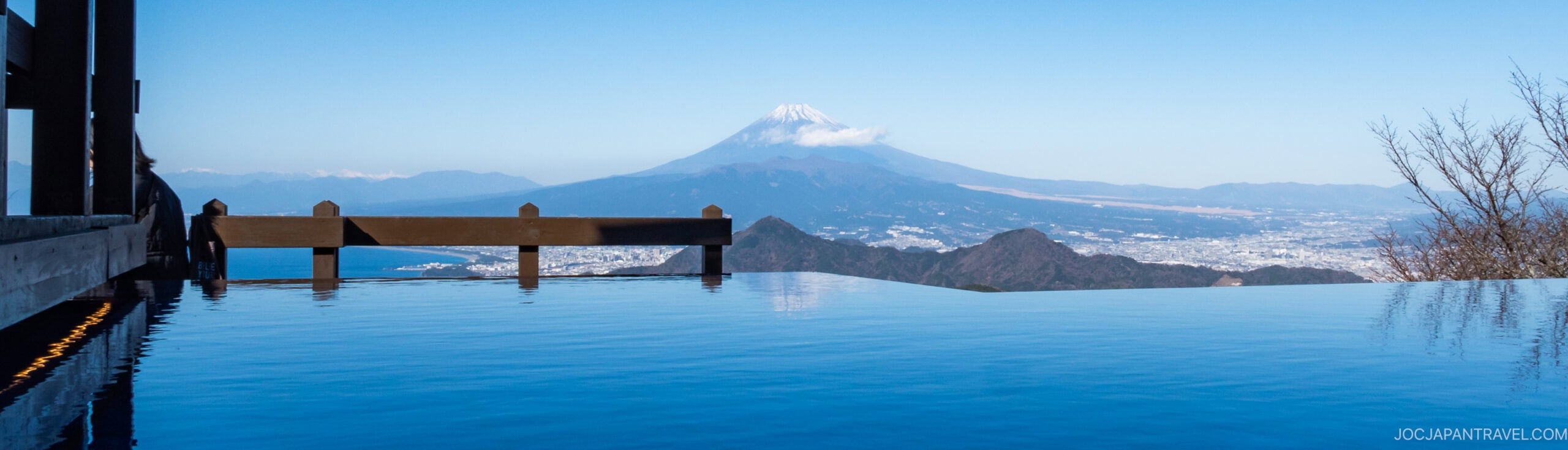 A serene infinity pool overlooks a vast landscape with Mount Fuji in the distance under a clear blue sky, reflecting the mountain and sky in its calm water.