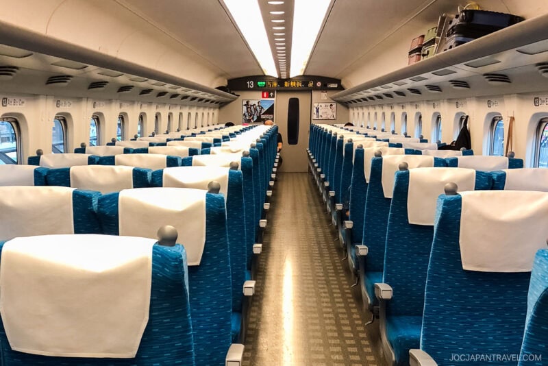 Interior of a modern Japanese bullet train (Shinkansen) with rows of empty blue seats, white headrest covers, overhead luggage racks, and a digital sign at the front—accessible with a Japan Rail Pass for seamless travel across Japan.