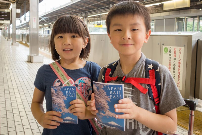 Two children stand on a train platform, smiling and holding their Japan Rail Pass booklets. With backpacks on and the station behind them, they appear excited to begin their adventure across Japan.