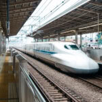 How to Purchase and Use Japan Rail Pass (JR Pass) – Updated for 2025 A white Shinkansen bullet train is stopped at a covered station platform in Japan, with overhead wires and signs visible. Passengers, some using a Japan Rail Pass, and vending machines can be seen on the platform.
