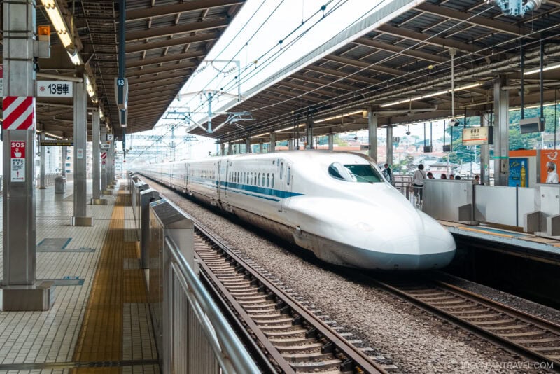 A white Shinkansen bullet train is stopped at a covered station platform in Japan, with overhead wires and signs visible. Passengers, some using a Japan Rail Pass, and vending machines can be seen on the platform.