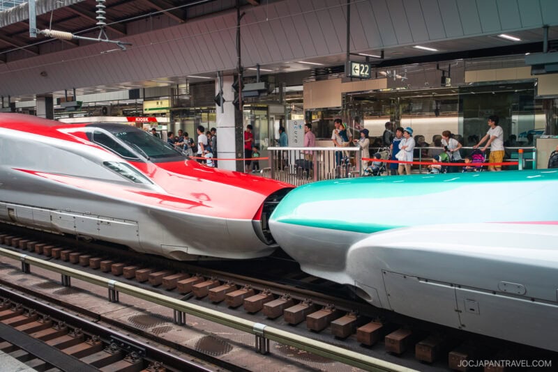 Two modern Shinkansen bullet trains, one red and one green, are coupled together at a station platform as passengers with Japan Rail Passes wait nearby. The covered station features visible signage above the sleek trains.