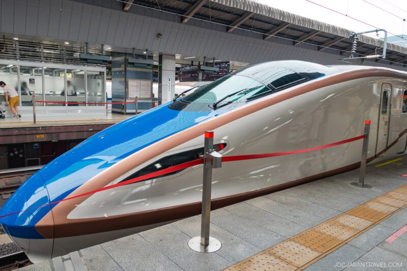 A sleek, modern Japanese bullet train with a blue and white streamlined nose is parked at a station platform under a covered roof, ready to whisk japan rail pass holders across the country in comfort and style.