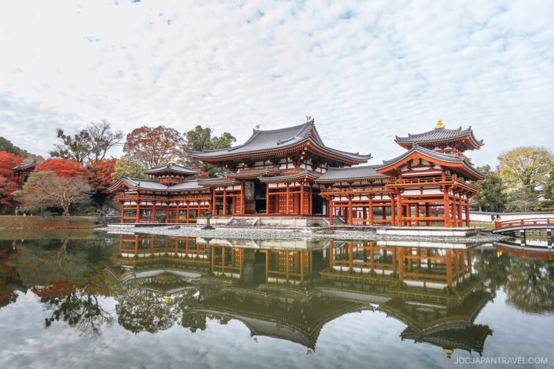 A traditional Japanese temple with ornate roofs and red pillars stands beside a calm pond, reflecting the building and autumn trees. Visitors respectfully observe Japanese Shrine and Temple Etiquette under the partly cloudy sky.
