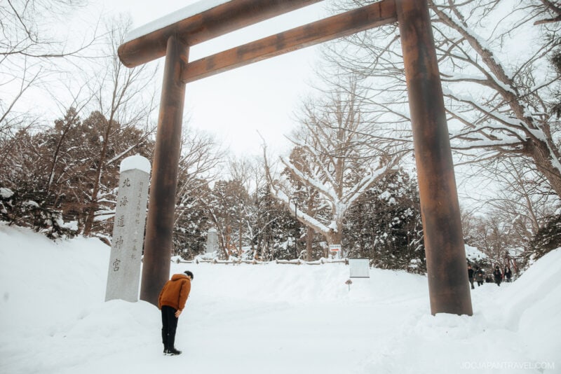 A person bows in front of a large wooden torii gate, surrounded by snow-covered trees and paths, peacefully observing Japanese Shrine and Temple Etiquette at a serene winter shrine.
