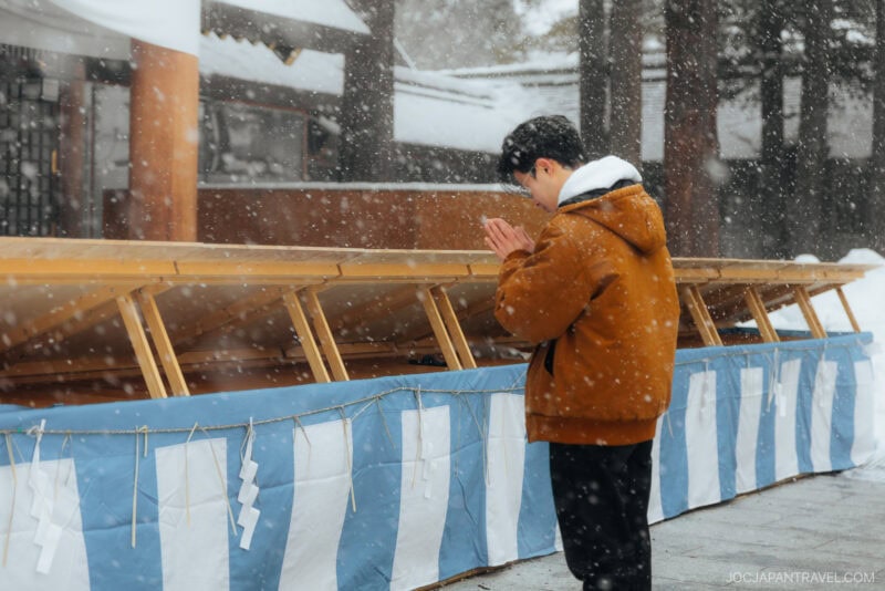 A person in a brown jacket stands with hands clasped in prayer at a snowy outdoor shrine, respectfully observing Japanese Shrine and Temple Etiquette, surrounded by gently falling snowflakes and wooden structures draped with blue and white cloth.