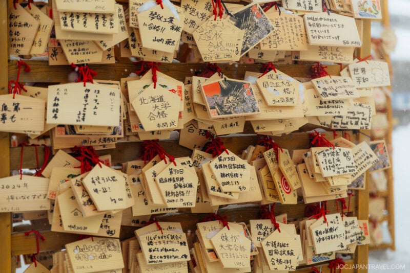 Wooden ema plaques hang on a display, each with handwritten wishes or prayers in Japanese, reflecting Japanese Shrine and Temple Etiquette, some adorned with illustrations or photos and tied with red strings.
