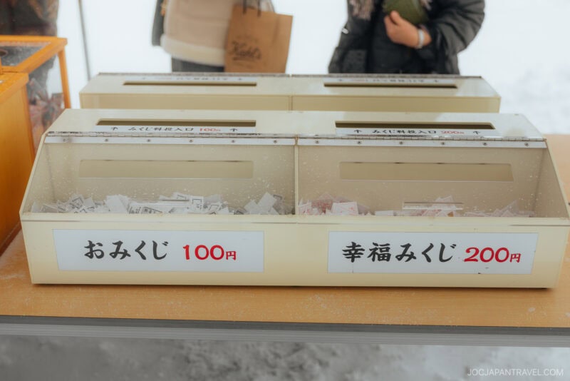Two boxes filled with omikuji (Japanese fortune-telling) slips, labeled 100 yen and 200 yen, rest on a snowy surface. In line with Japanese Shrine and Temple Etiquette, people wait in the background, bags in hand.