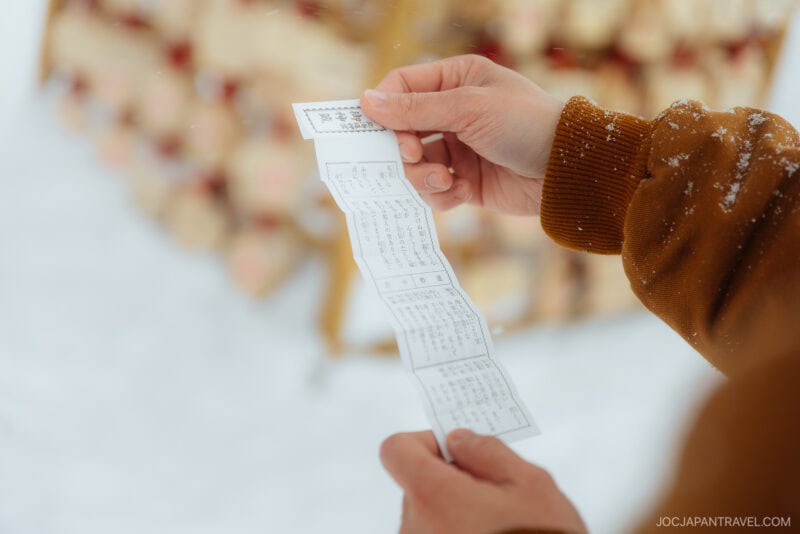 A person holding and reading a Japanese omikuji fortune slip outdoors in snowy surroundings, observing Japanese Shrine and Temple Etiquette, with wooden plaques blurred in the background.
