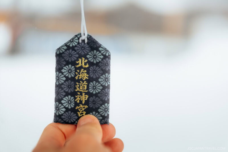 A hand holds a dark blue Japanese omamori (amulet) with gold kanji characters and a white cord, reflecting Japanese Shrine and Temple Etiquette, against a blurred snowy background.
