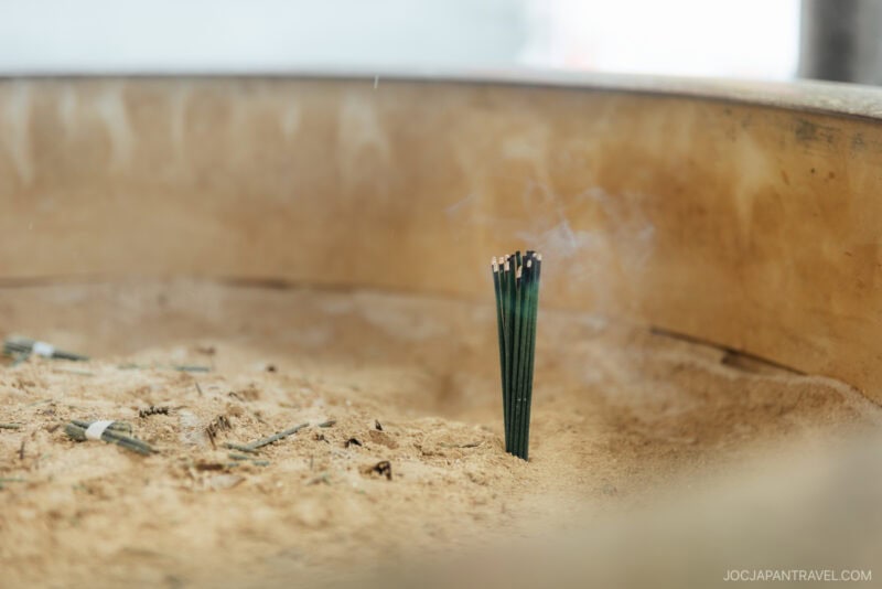 A small bunch of lit incense sticks stands upright in a bed of sand, with smoke rising gently—echoing Japanese Shrine and Temple Etiquette. The blurred background draws focus to the ritual’s serene beauty.