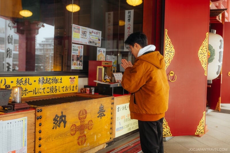 A person in an orange jacket stands in prayer with hands together at a donation box, observing Japanese Shrine and Temple Etiquette outside a traditional temple decorated with red pillars and gold details.
