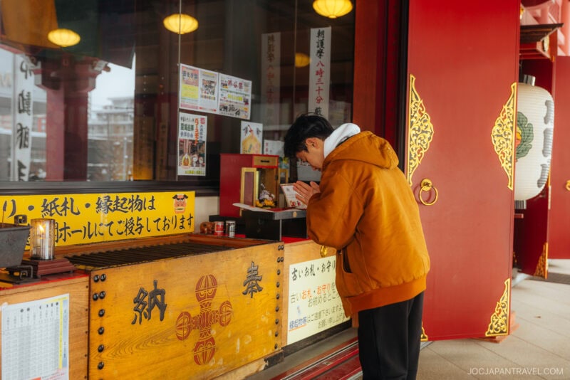 A person in an orange jacket stands at a wooden offering box outside a Japanese temple, bowing their head in prayer. The scene, rich with red pillars and gold details, reflects traditional Japanese Shrine and Temple Etiquette.