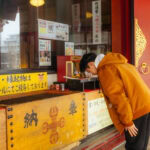 A Beginner’s Guide to Japanese Shrine and Temple Etiquette A person in an orange jacket bows at a wooden offertory box in front of a colorful shrine, practicing Japanese Shrine and Temple Etiquette, with signs and ornate red and gold details visible around the entrance.