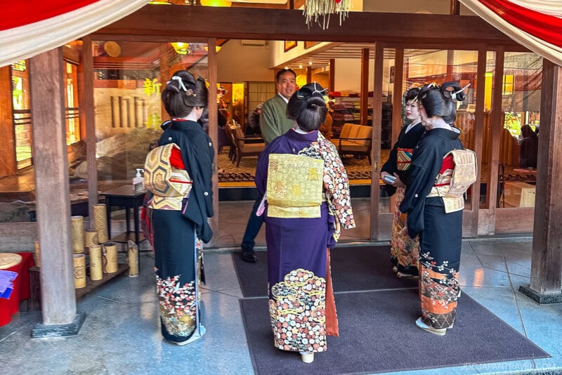 Four women in traditional Japanese kimonos with ornate obi sashes stand at the entrance of a building, talking to a man welcoming them inside—a graceful moment that reflects customs found in any Japanese Etiquette Guide.