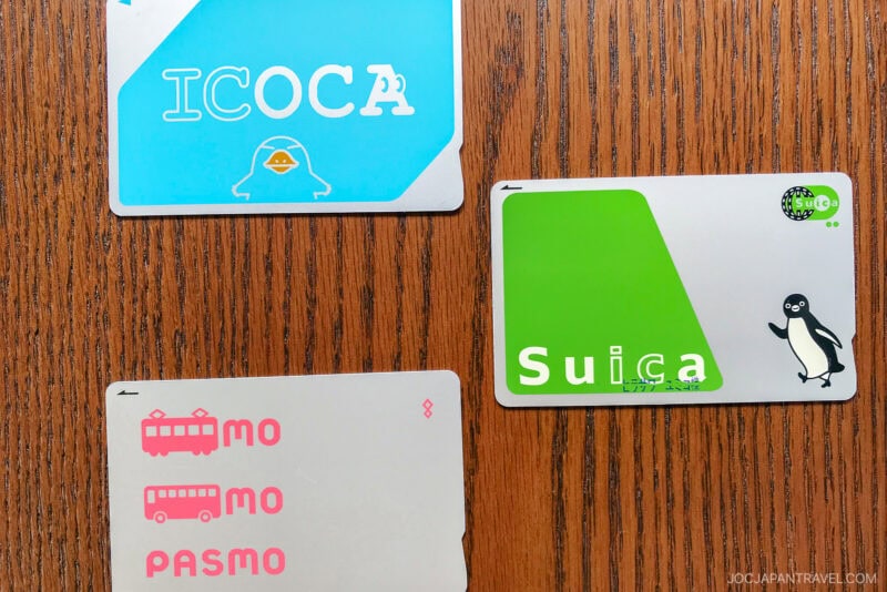 Three Japanese transportation cards—ICOCA (blue), Suica (green with a penguin), and PASMO (white with pink text)—are arranged on a wooden surface, offering a glimpse into travel convenience and Japanese etiquette guide essentials.