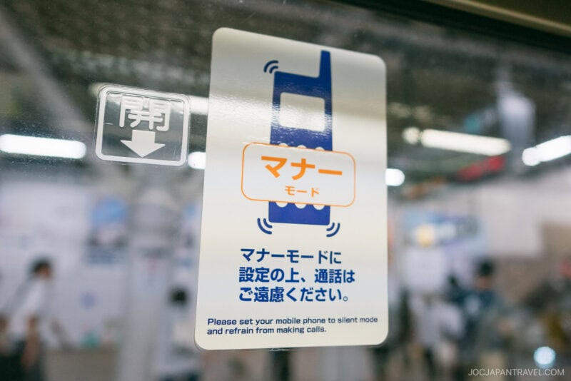 A sign on a glass window in a public place, styled like a Japanese Etiquette Guide, instructs people in both Japanese and English to set mobile phones to silent mode and avoid calls. Blurry figures and objects can be seen in the background.