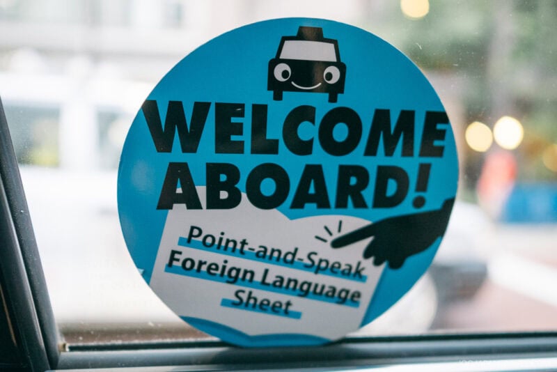 A blue circular sign on glass reads, WELCOME ABOARD! Point-and-Speak Foreign Language Sheet, featuring a cartoon car and a pointing hand—perfect for travelers seeking a handy Japanese Etiquette Guide.