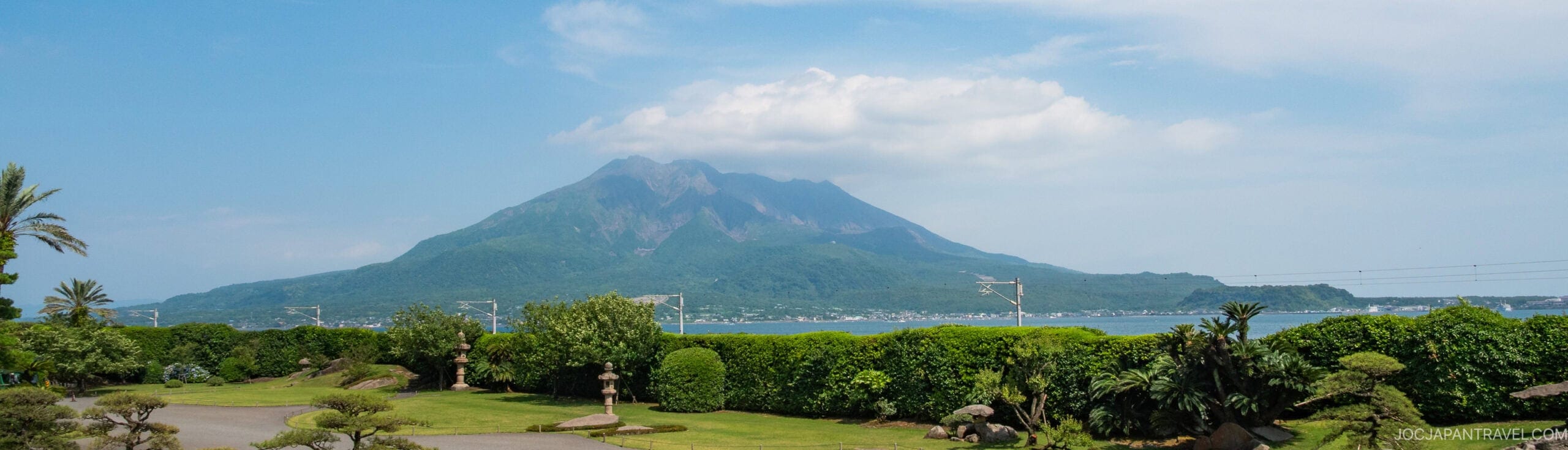 A lush green garden with palm trees and shrubs overlooks a calm bay, with a large, cloud-topped volcanic mountain rising in the background under a blue sky.