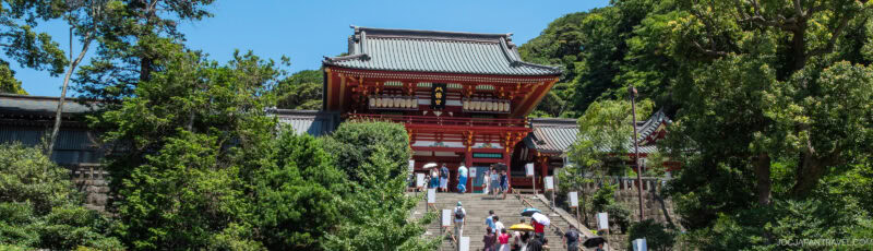 A group of people walk up stone steps toward a large, traditional red Japanese shrine surrounded by lush green trees under a clear blue sky.