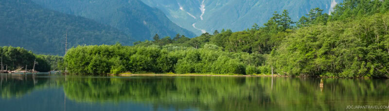 A calm lake reflects lush green trees and dense forest, with tall mountains rising in the background under a clear sky.