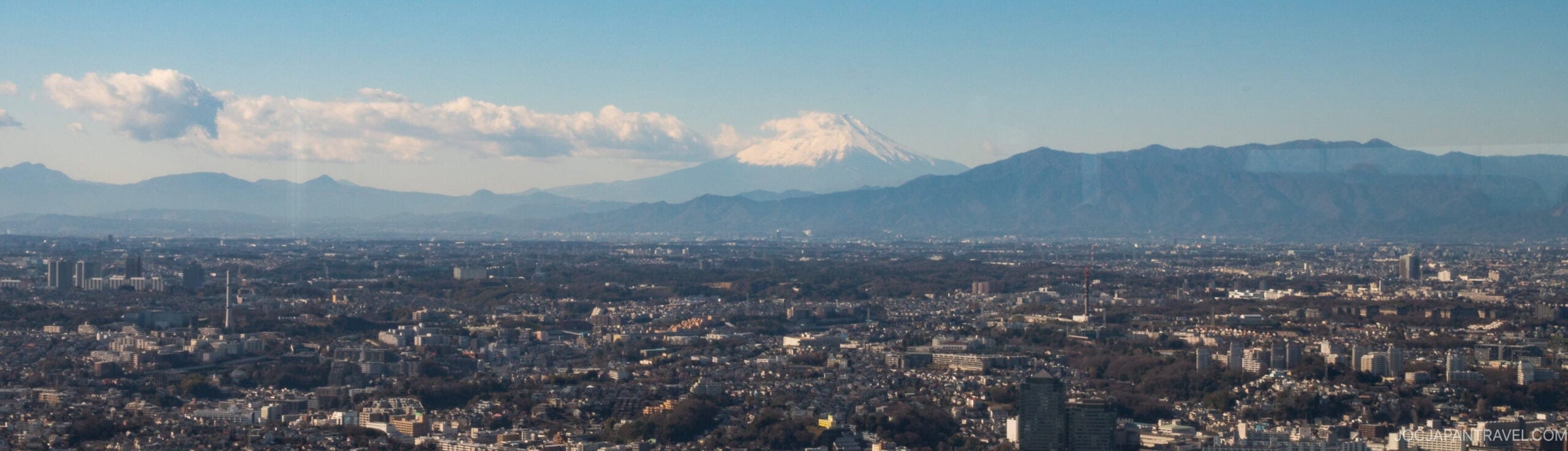 Panoramic view of a city with Mount Fuji in the background, its snow-capped peak rising above the surrounding mountains under a clear blue sky.