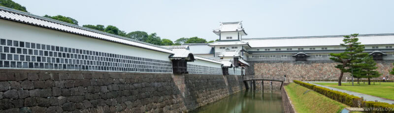 A traditional Japanese castle with white walls, a stone base, and tiled roof sits behind a moat and wooden bridge, surrounded by trimmed grass and trees under a clear sky.