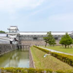 Kanazawa Castle Park (金沢城公園) – A Gateway to Feudal Japan Kanazawa Castle Park features a traditional Japanese castle with white walls and tiled roofs, set behind a stone moat and surrounded by green lawns, trees, and neatly trimmed hedges under a clear sky.