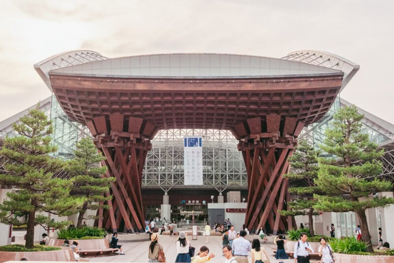 The large wooden gate with unique curved supports marks the entrance of Kanazawa Station&rsquo;s modern glass building, surrounded by trees and people walking in front. The sky is clear and bright.